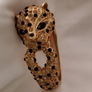 Puede incluir: Brazalete dorado con diseño de cabeza de leopardo. El brazalete está adornado con pedrería ámbar y negra, creando un efecto brillante. Los ojos del leopardo son negros, y el diseño general es detallado y elegante.