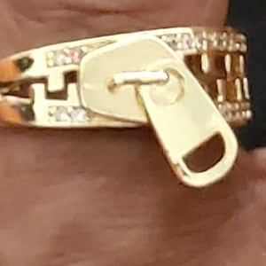 May include: Gold-toned ring with a zipper design. The ring features a zipper pull and is embellished with small, clear gemstones. The ring is a unique piece of jewelry.