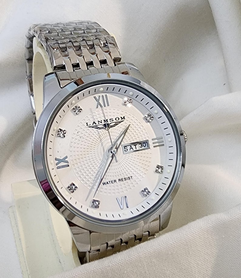 Op de afbeelding: Een zilverkleurig polshorloge met een witte wijzerplaat met Romeinse cijfers, diamantachtige markeringen en de dag van de week. Het horloge heeft een getextureerde wijzerplaat en een roestvrijstalen band. De wijzerplaat leest "LANMSOM" en "WATER RESIST".
