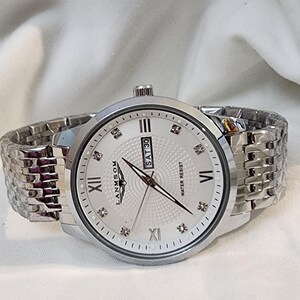 Op de afbeelding: Zilverkleurig polshorloge met een lichtgekleurde wijzerplaat, Romeinse cijfers en diamantachtige markeringen. De horloge heeft een datumweergave en een metalen schakelband. De merknaam "LANMSOM" is zichtbaar. Waterbestendig.