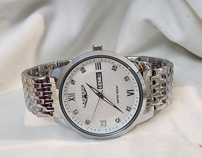 Op de afbeelding: Zilverkleurig polshorloge met een lichtgekleurde wijzerplaat, Romeinse cijfers en diamantachtige markeringen. De horloge heeft een datumweergave en een metalen schakelband. De merknaam "LANMSOM" is zichtbaar. Waterbestendig.