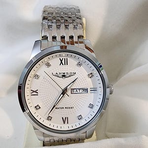 Op de afbeelding: Een zilverkleurig polshorloge met een witte wijzerplaat met Romeinse cijfers en diamantachtige markeringen. De wijzerplaat toont de dag en datum, met de merknaam "LANMSOM". Het horloge heeft een roestvrijstalen band.