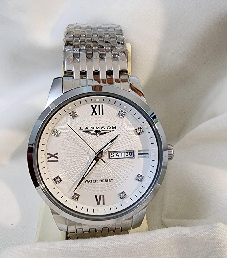 Op de afbeelding: Een zilverkleurig polshorloge met een witte wijzerplaat met Romeinse cijfers en diamantachtige markeringen. De wijzerplaat toont de dag en datum, met de merknaam "LANMSOM". Het horloge heeft een roestvrijstalen band.