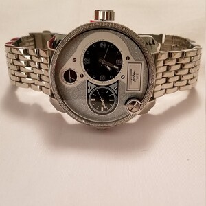May include: A silver watch with a diamond-encrusted bezel and a multi-layered dial. The watch has a silver metal band and a black face with white accents. The watch face features the text "Techno Pave".