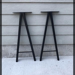 Puede incluir: Dos patas de mesa de metal negro en forma de A con un travesaño de apoyo. Las patas están sobre una superficie de hormigón.
