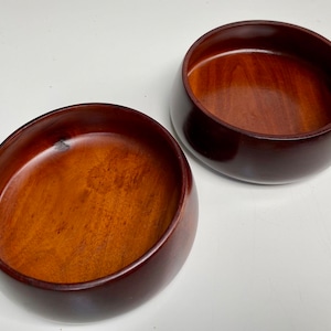 May include: Two wooden bowls, one slightly larger than the other, are shown on a white surface. Both bowls are a rich brown color and have a smooth, polished finish.