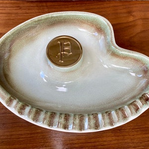 May include: A green and brown ceramic ashtray with a gold metal 'P' in the center.