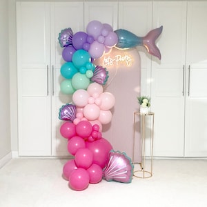 Może przedstawiać: Łuk balonowy w stylu syreny z pastelowymi różowymi, fioletowymi, niebieskimi i zielonymi balonami. Łuk jest ozdobiony balonem w kształcie ogona syreny i neonowym napisem "Let's Party".