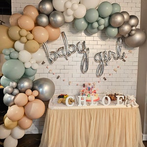 May include: A baby shower party decoration with a balloon arch in shades of white, silver, sage green, and peach. The arch is decorated with the words "baby girl" in silver foil letters. The table is decorated with a tulle tablecloth and the name "Chloe" spelled out in white letters.