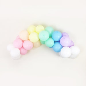 May include: A rainbow-shaped arrangement of small, pastel-coloured balls. The colours include white, pink, peach, yellow, mint green, light blue, and lilac. The balls are arranged on a plain, off-white background.
