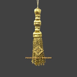 May include: Gold tassel with a diamond-shaped design featuring the letter "P" in the center. The tassel is made of braided cord and has a decorative top. The text "PASAMANERIAS Y BORDADOS" is visible below the tassel.