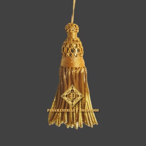 Könnte beinhalten: Ein goldener Quaste mit einem dekorativen Oberteil und einem rautenförmigen Emblem mit dem Buchstaben "P" in der Mitte. Die Quaste ist mit "PASAMANERIAS Y BORDADOS" beschriftet.