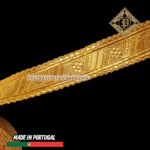 May include: Gold-colored decorative trim with intricate floral and geometric patterns. The trim has a textured surface and is labeled "PASAMANERIAS Y BORDADOS" and "MADE IN PORTUGAL". The trim is against a black background.