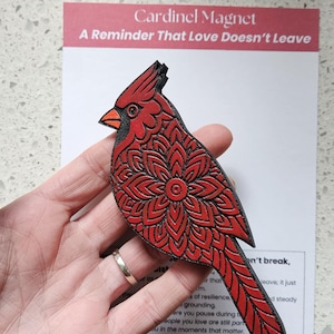 May include: A red cardinal magnet with detailed floral patterns and black outlines. The magnet reads 'A Reminder That Love Doesn't Leave'. The cardinal has an orange beak. The magnet is held in a hand.