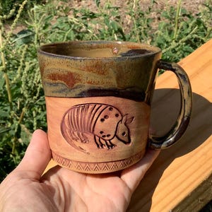 May include: A ceramic mug featuring a carved armadillo design on the front. The mug has a two-tone finish with a smooth, dark brown glazed upper section and a natural, unglazed terracotta-colored lower section with a geometric border pattern.
