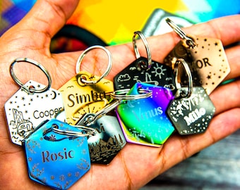 May include: A collection of seven hexagonal metal keychains with different designs and names engraved on them. The keychains are in various colors, including silver, gold, rainbow, and blue. The names on the keychains are "Simba", "Venus", "Milo", "Cooper", "Rosie", and "HOR".