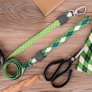May include: Two green and white patterned dog leashes with black metal clips and a black metal ring. One leash has a white polka dot pattern and the other has a green and white plaid pattern. The leashes are on a wooden surface with a pair of black scissors.