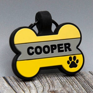 以下が含まれることがあります： 黄色と灰色の骨の形をした犬のタグで、"COOPER"という名前が印刷されています。タグの右下には黒い足跡があります。