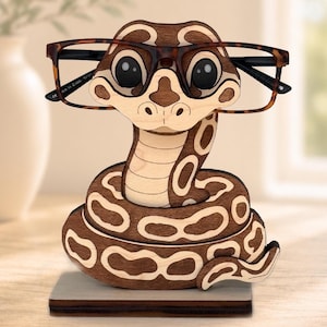 May include: A wooden eyeglass holder shaped like a coiled snake with a brown and tan patterned body. The snake has large, dark eyes and is designed to hold a pair of brown tortoiseshell-patterned glasses across its head on a flat base.