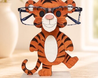 May include: A wooden tiger-shaped eyeglass holder featuring orange fur with black stripes, a white belly, and a pink nose. The tiger stands on a rectangular base and holds a pair of blue and grey patterned eyeglasses across its face.