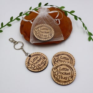 May include: Four wooden keychain tags with the text "Little Pocket Hug" and a heart design. The tags are on a white surface with a small green plant in the background.
