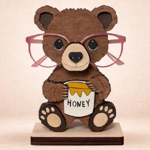 Może przedstawiać: Drewniany uchwyt na okulary w kształcie brązowego misia. Miś ma różowe okulary i trzyma słoik miodu. Na słoiku wydrukowano napis "HONEY". Uchwyt stoi na prostokątnej drewnianej podstawie.