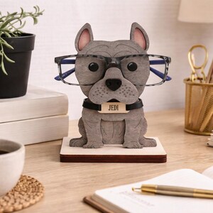 May include: A gray bulldog-shaped glasses holder with a black collar and a name tag that reads "Jedi." The holder is wearing glasses and sits on a wooden base. A coffee cup, plant, and desk supplies are in the background.