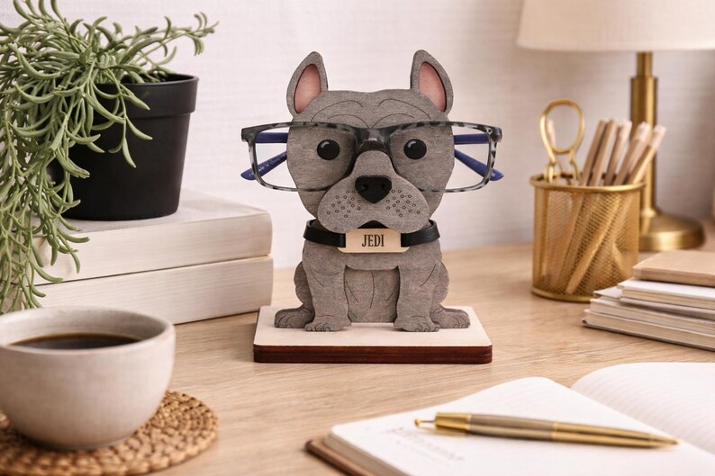 May include: A gray bulldog-shaped glasses holder with a black collar and a name tag that reads "Jedi." The holder is wearing glasses and sits on a wooden base. A coffee cup, plant, and desk supplies are in the background.