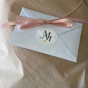 May include: A small, silver envelope tied with a light pink ribbon. A round sticker with the letters "MM" and a small heart is on the envelope. The envelope is on a brown, textured surface.