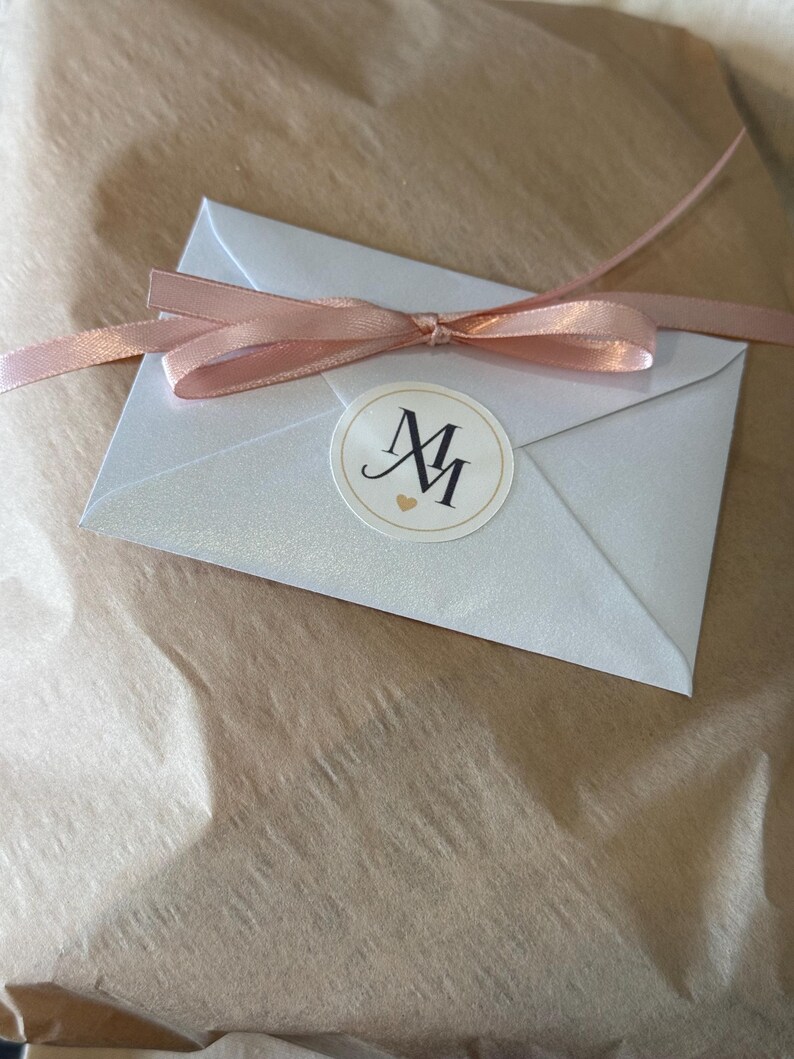 May include: A small, silver envelope tied with a light pink ribbon. A round sticker with the letters "MM" and a small heart is on the envelope. The envelope is on a brown, textured surface.