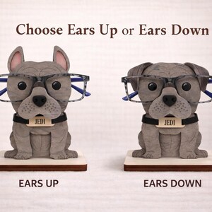 May include: Two gray bulldog-shaped eyeglass holders, one with ears up and the other with ears down, each wearing glasses. The holders have a black collar with the name "JEDI" on a tag. The text "Choose Ears Up or Ears Down" is above the dogs.