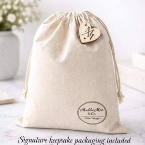 May include: A beige drawstring bag with a wooden heart-shaped tag. The tag has an initial and is attached to the bag. The bag has the text "Maddie Mae & Co." and "Little Things" printed on it. The bag is made of a soft, natural fabric.