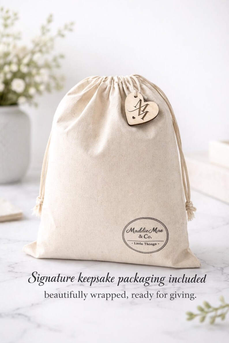 May include: A beige drawstring bag with a wooden heart-shaped tag. The tag has an initial and is attached to the bag. The bag has the text "Maddie Mae & Co." and "Little Things" printed on it. The bag is made of a soft, natural fabric.