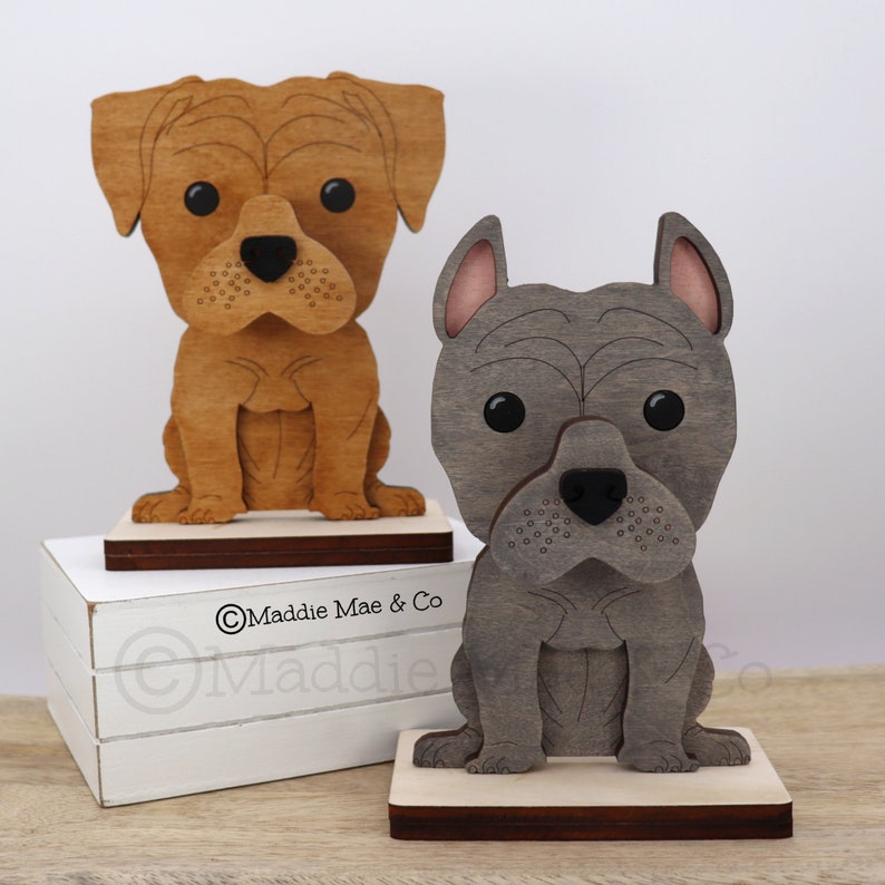 May include: Two wooden dog figurines. One is light brown, the other is gray. Both are sitting on a wooden base. The light brown dog has a black nose and large black eyes. The gray dog has pink inner ears and a black nose.