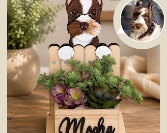 May include: A wooden planter box featuring a cutout of a brown and white dog peeking over a fence. The front of the light-colored wooden box displays the name Mocha in black script lettering. The planter contains green and purple artificial succulents.