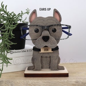 May include: A wooden eyeglasses holder shaped like a gray bulldog wearing glasses. The dog has a black collar with a name tag that reads "JEDI". The holder is on a wooden base. The text "EARS UP" is above the dog's head.