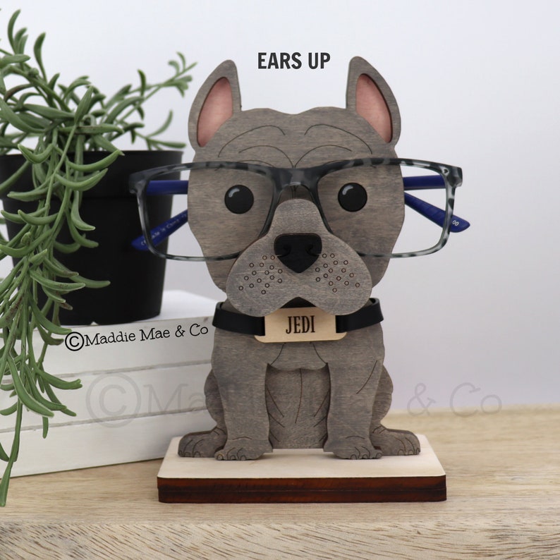 May include: A wooden eyeglasses holder shaped like a gray bulldog wearing glasses. The dog has a black collar with a name tag that reads "JEDI". The holder is on a wooden base. The text "EARS UP" is above the dog's head.
