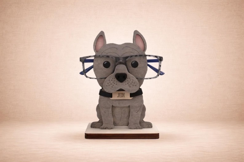 May include: A gray bulldog-shaped eyeglass holder with black-rimmed glasses. The dog wears a black collar with a tag that reads "JEDI". The holder sits on a white and brown rectangular base. The background is a solid light brown.
