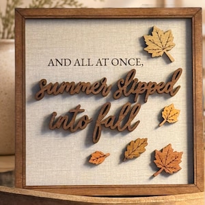 Puede incluir: Un letrero cuadrado de madera con un fondo de tela gris claro que presenta el texto "AND ALL AT ONCE, summer slipped into fall" en letras de color marrón oscuro. El letrero está decorado con cinco recortes de hojas de madera.