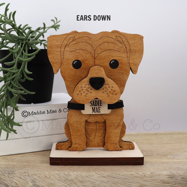 May include: Wooden dog figurine with the name "Sadie Mae" on a tag. The dog is light brown with black eyes and a black collar. The text "EARS DOWN" is above the dog's head.