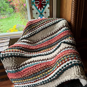 May include: A hand-knitted blanket with a chunky texture, featuring stripes of beige, rust, teal, and brown. The blanket's design creates a cosy and inviting look. Ideal for a bed or sofa, it adds warmth and style.