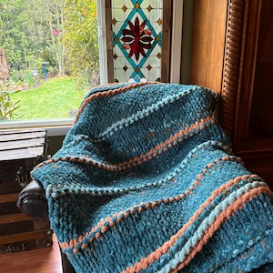 May include: A chunky knit throw blanket featuring a deep teal base with horizontal stripes in peach and light blue. The blanket has a thick, textured yarn construction with raised bobble-like details running across the width of the fabric.