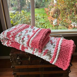 May include: A white ceramic mug with a black handle and the text "But First, Coffee" in a bold, black font. The mug is set against a blurred background, suggesting a cozy setting.
