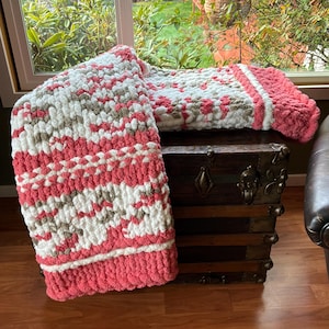 May include: A handmade, chunky knit blanket in shades of coral, white, and gray. The blanket is folded over a wooden trunk, showcasing its textured pattern and thick, cozy appearance. The blanket's edges are a solid coral color.