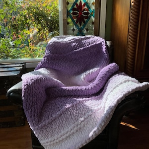 May include: A chunky knit blanket in shades of purple and white is draped over a black leather armchair. The blanket's texture is visible, with a gradient effect from dark purple to white. The scene is illuminated by natural light.