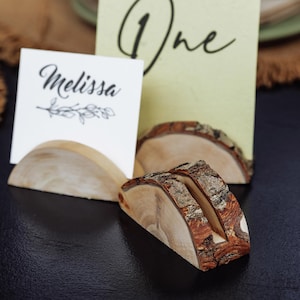 May include: Wooden place card holders with a natural bark edge. One holder displays the name "Melissa" and another displays the number "One". The holders are on a black surface, ideal for weddings or events.