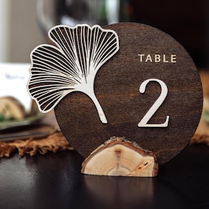 May include: Wooden table number 2, with a laser-cut ginkgo leaf design. The number is on a dark brown circular base, with the word "TABLE" above it. The base is supported by a small wooden slice.