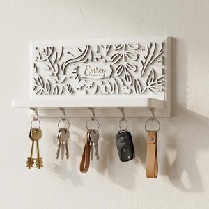 May include: A white wall-mounted key holder featuring a rectangular shelf and five silver metal hooks. The upper panel displays a laser-cut floral pattern and the text "Emrey FAMILY" in the centre. Five sets of keys hang from the hooks below.