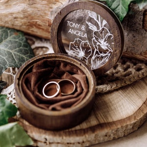 May include: A wooden ring box with a glass lid. The lid has a floral design and the text "Tony & Angela". The box is open and two gold wedding bands are inside on a brown velvet cushion.