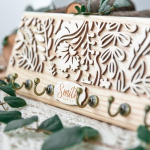 May include: Wooden key holder with a floral design and five hooks. The key holder has a sign that reads "Smith Family".